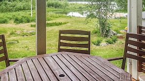 Une terrasse extérieure avec une table ronde en bois et deux chaises en bois. La terrasse donne sur une zone herbeuse, avec des arbres et une étendue d’eau en arrière-plan.