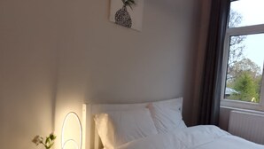 Un lit bien fait, avec du linge de lit blanc, une table de chevet avec une lampe et un vase de fleurs, et une fenêtre donnant sur des arbres.