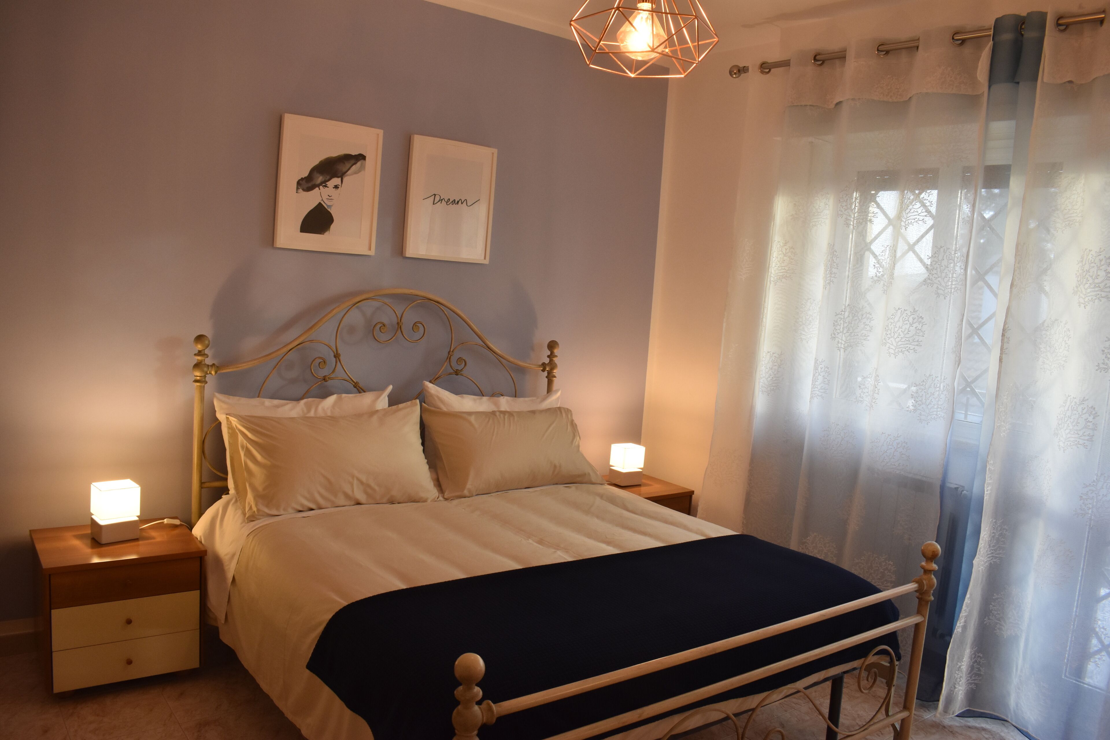 A bedroom with a bed, two bedside tables, a nightstand, a wall-mounted light, and a window with sheer curtains.
