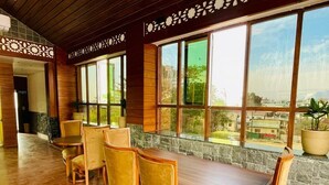 A restaurant with a long wooden table and chairs, large windows with a view of the city, and a stone wall. - Hotel Suryadev (Palampur)