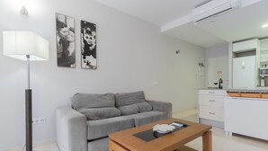 A modern living room with a grey sofa, a wooden coffee table, and a floor lamp. There is a kitchen area with a white refrigerator and a microwave. - Apartamento Premium un Dormitorio con Terraza (Fuengirola)