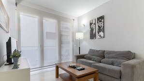 A modern living room with a grey sofa, a wooden coffee table, and a floor lamp. There is a large window with blinds, a potted plant, and a flat-screen TV on a stand. - Apartamento Premium un Dormitorio con Terraza (Fuengirola)