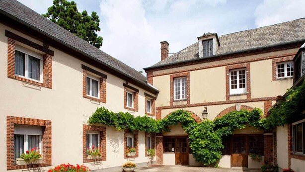 Une cour bordée de bâtiments à deux étages, aux façades en brique et en crépi, et agrémentée de plantes en pot.