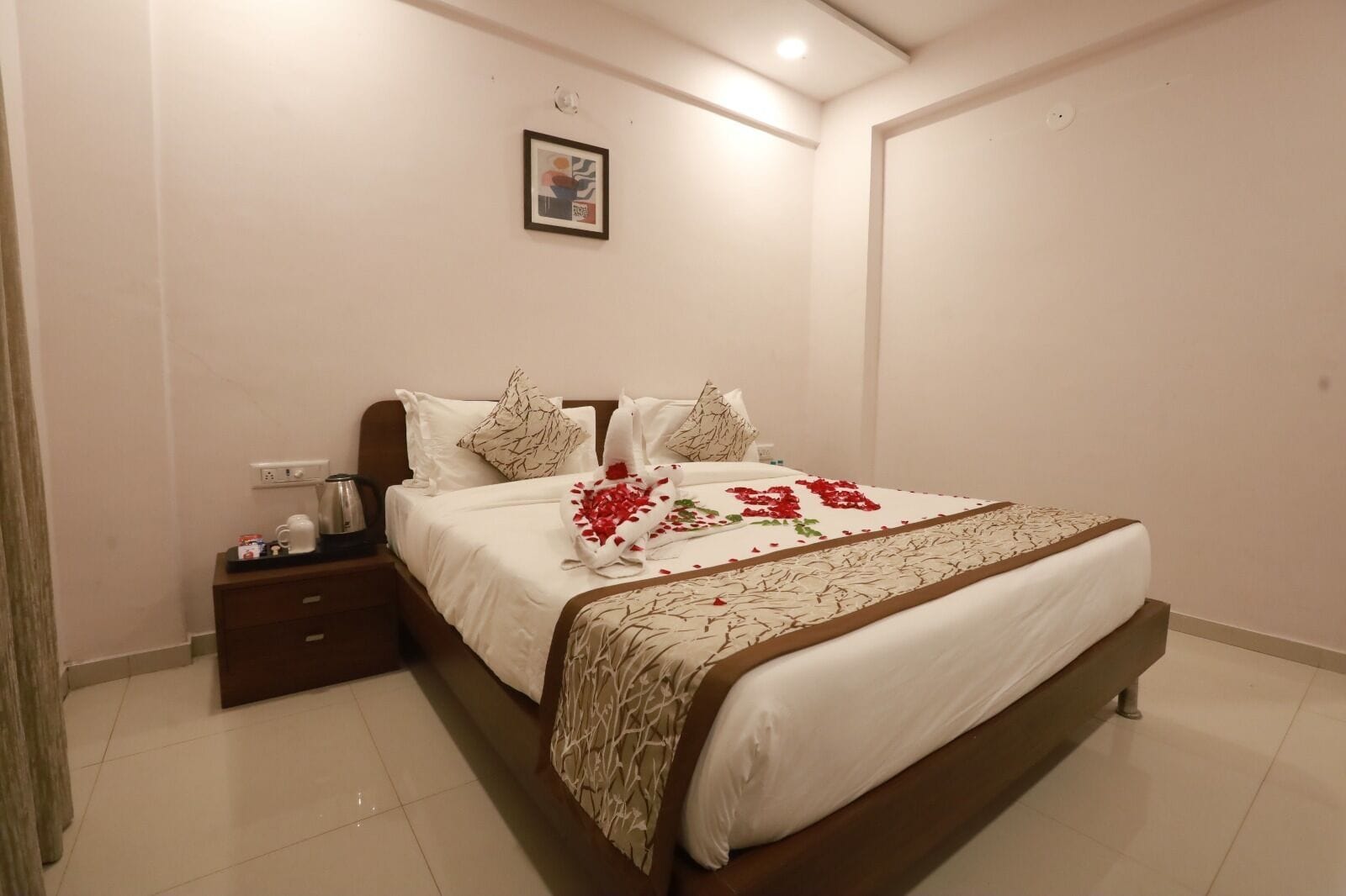 A neatly made bed with a floral bedspread, a bedside table with a kettle, and a framed picture on the wall.