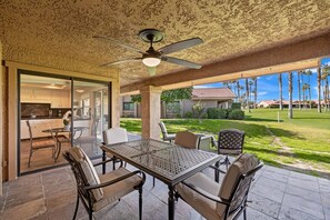 Um pátio externo com mesa e cadeiras, um ventilador de teto e vista para uma área residencial com palmeiras e uma casa.