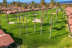 Um campo de golfe com palmeiras, um green bem cuidado e um edifício com telhado vermelho ao primeiro plano.
