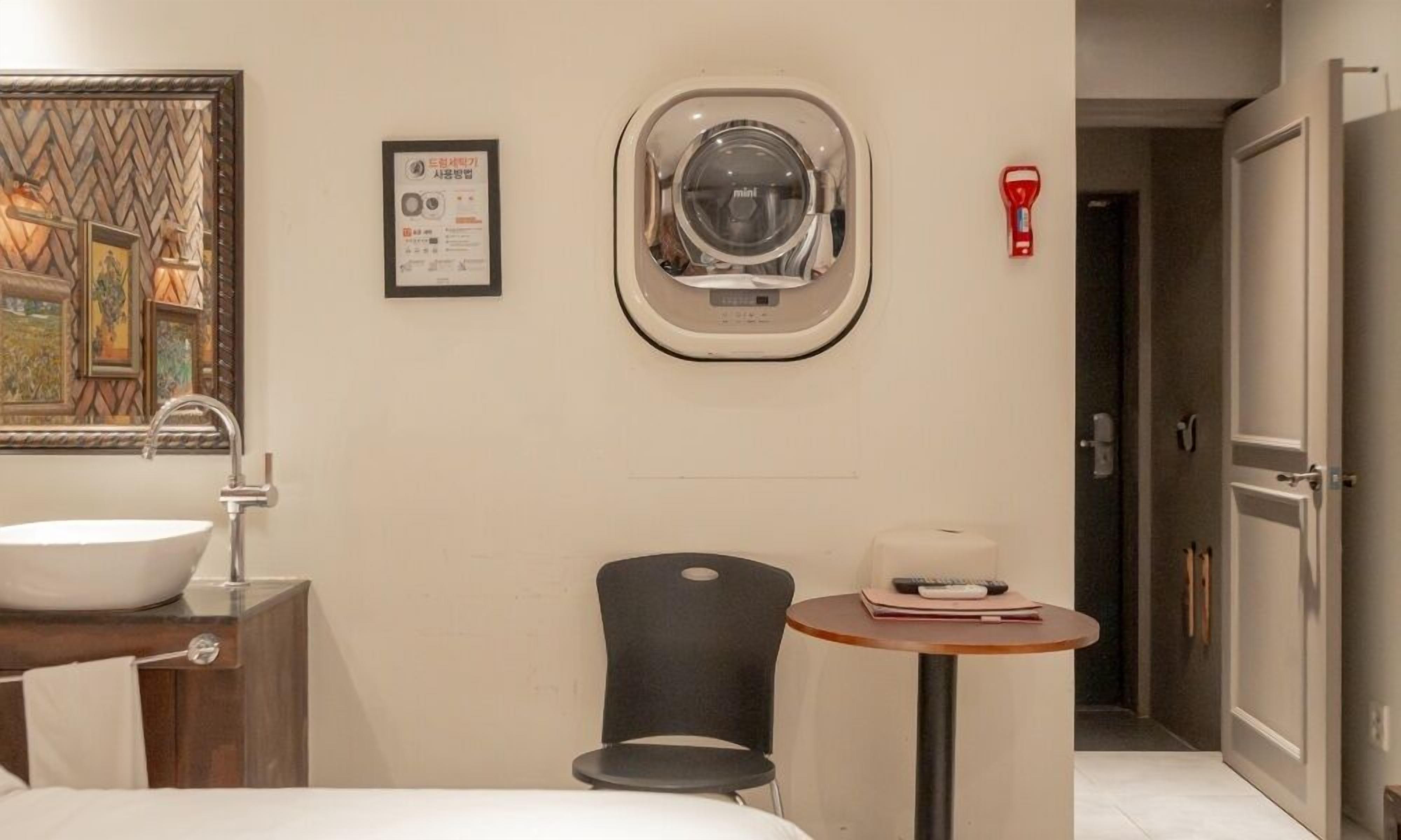 A hotel bathroom with a large sink, a mirror, a small window, a chair, and a table with a book and remote control.
