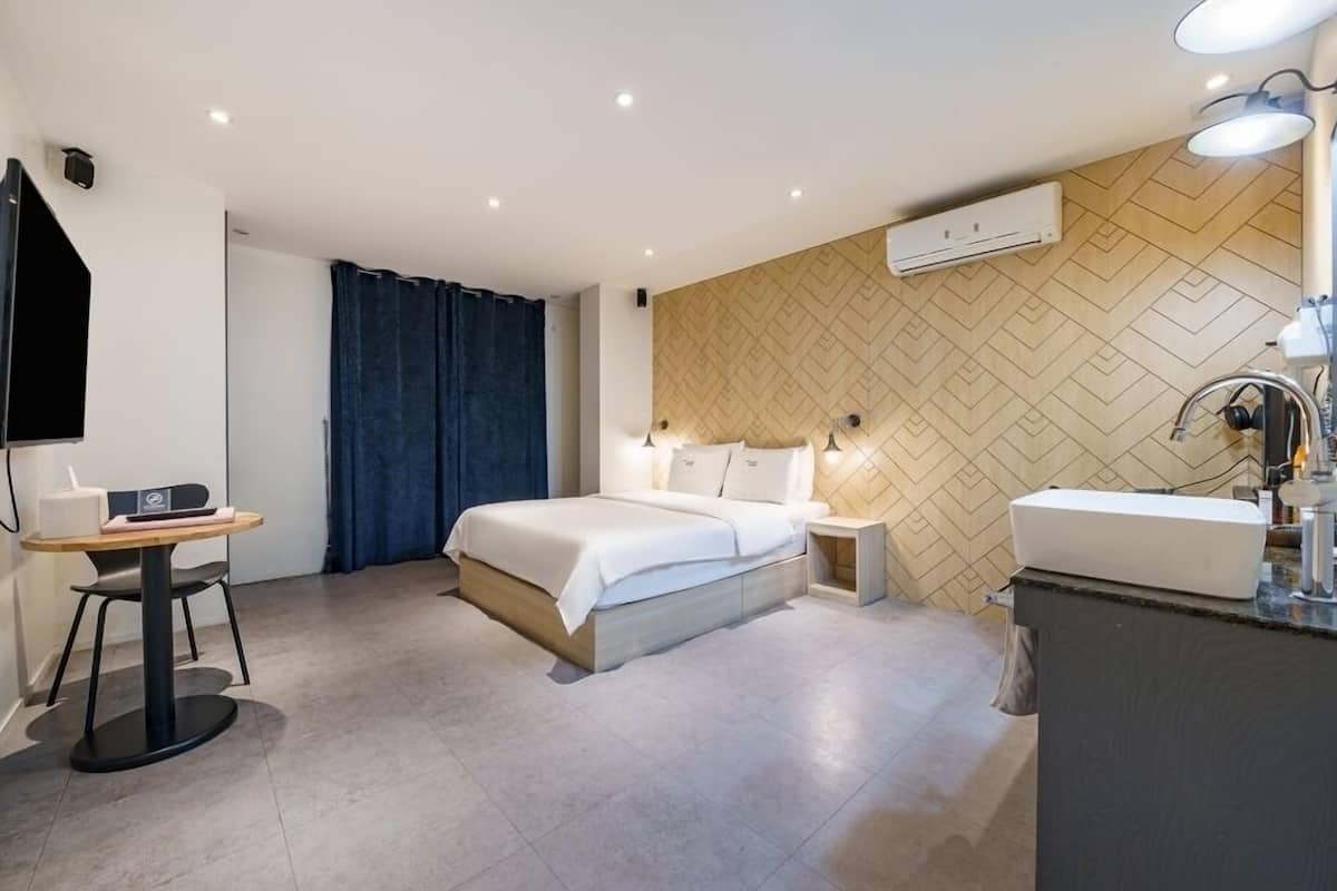 A modern hotel room with a bed, bedside table, desk, and a bathroom with a sink and mirror.