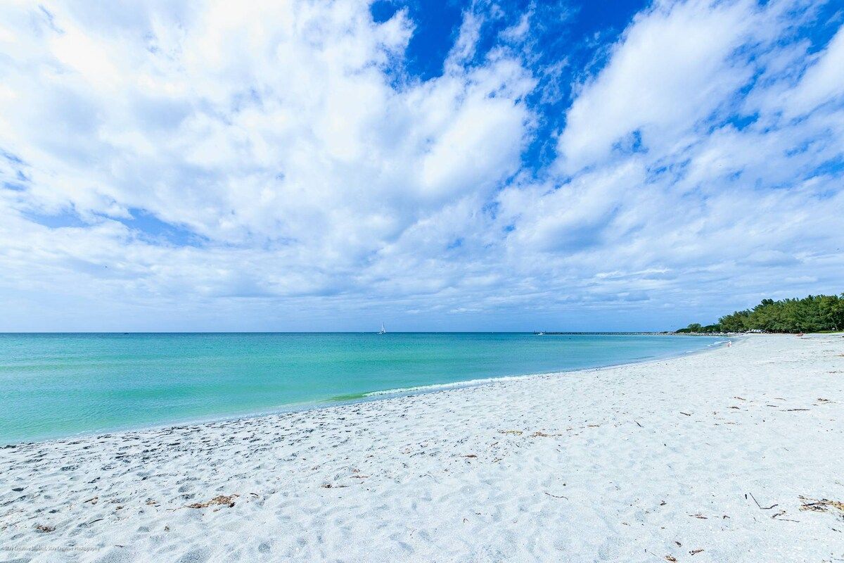 A beach with white sand and clear turquoise water under a partly cloudy sky.