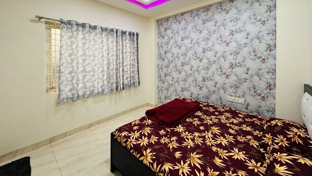 A bedroom with a bed, a ceiling fan, a window with curtains, and a floral wallpaper on the wall.