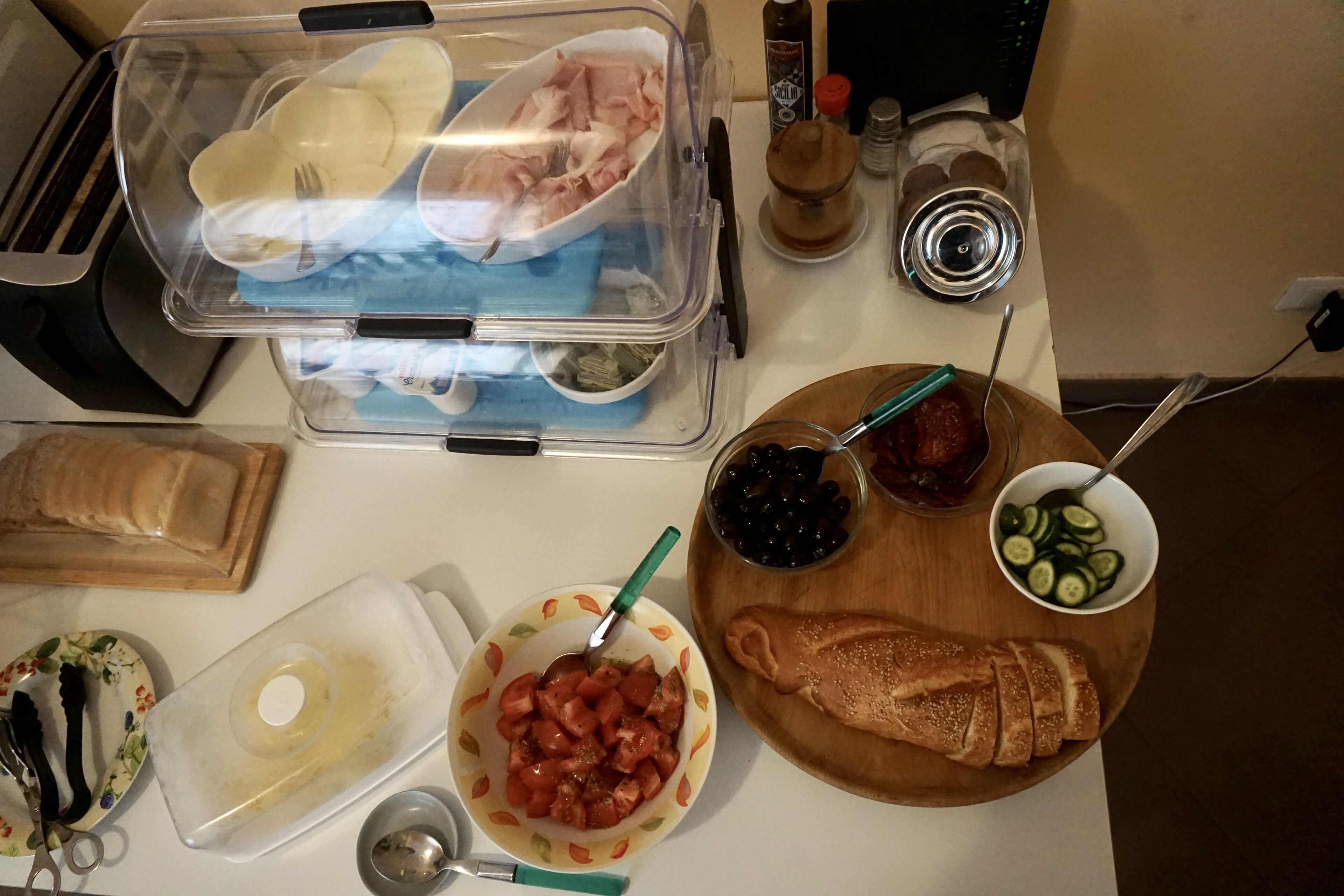 Un plan de travail de cuisine avec divers aliments et ustensiles, notamment une miche de pain, un bol de tomates et un récipient d’olives.