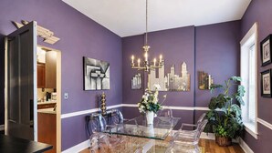 A dining area with a glass table and chairs, a chandelier, and a cityscape painting on the wall. - Charming 3BR | Highly Walkable | Free Parking! (Louisville)