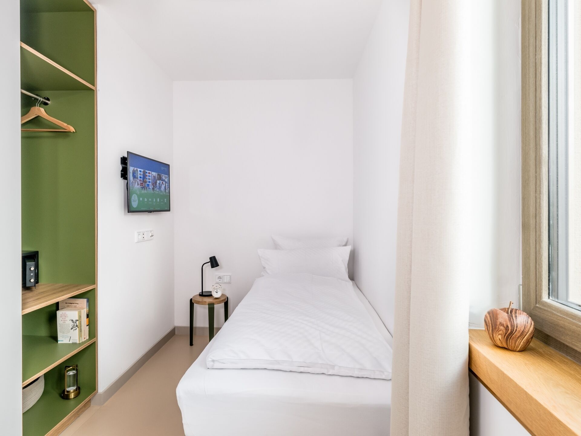 A small, neatly organized bedroom with a bed, a TV mounted on the wall, a bedside table with a lamp, and a window with a view.