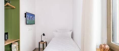 A small, neatly organized bedroom with a bed, a TV mounted on the wall, a bedside table with a lamp, and a window with a view.