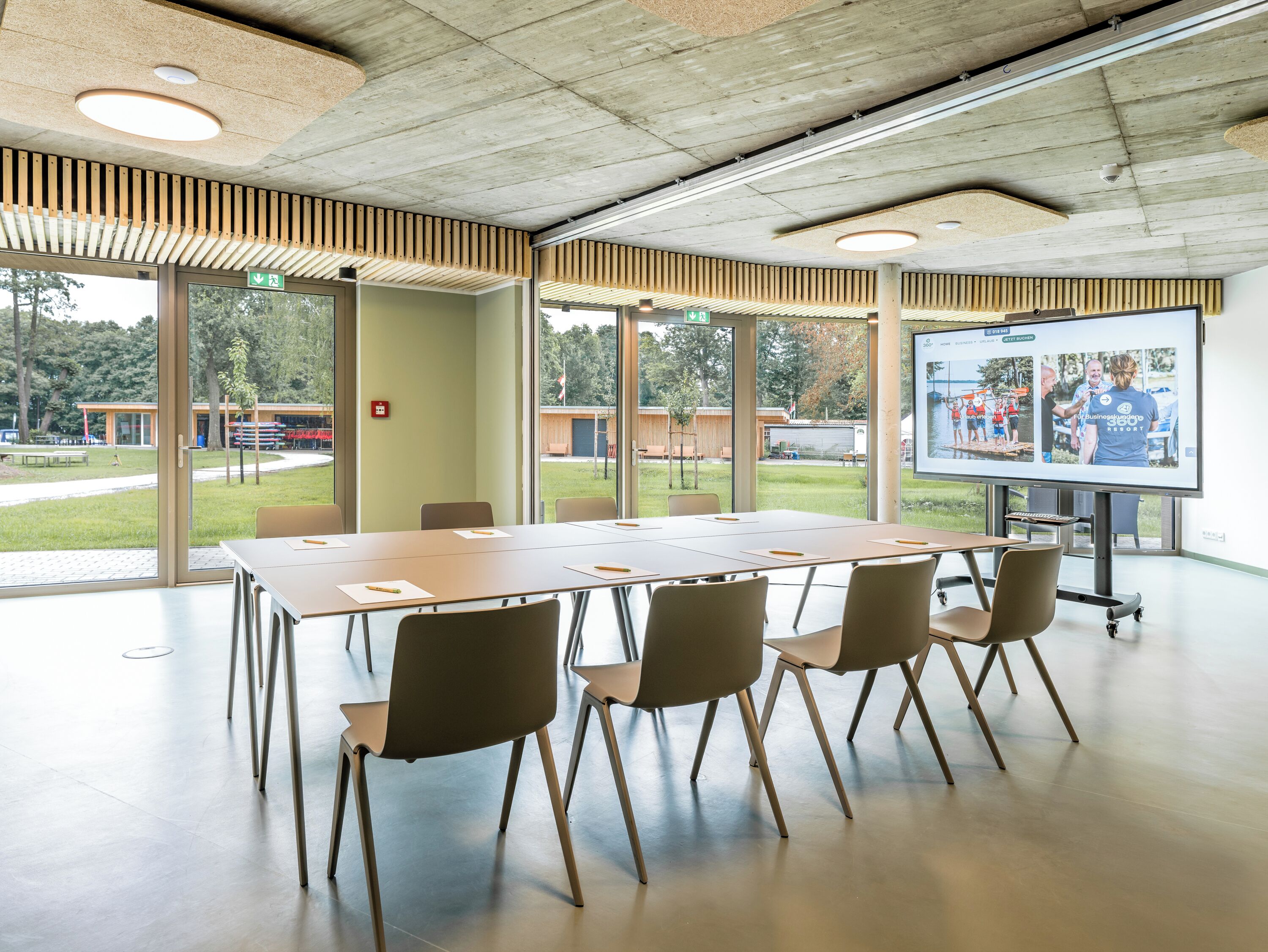 Ein Konferenzraum mit einem langen Tisch, Stühlen, einer Projektionsleinwand und großen Fenstern mit Blick ins Freie.