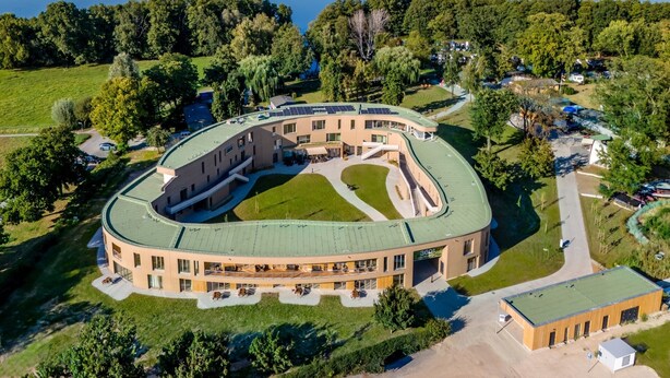 A large, circular building with a green roof, surrounded by a lush green landscape and a lake. - 360° Apartmenthotel - by teamgeist (Heidesee)