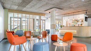 A modern lounge area with various seating options including chairs and ottomans, a reception desk, and large windows providing natural light. - 360° Apartmenthotel - by teamgeist (Heidesee)