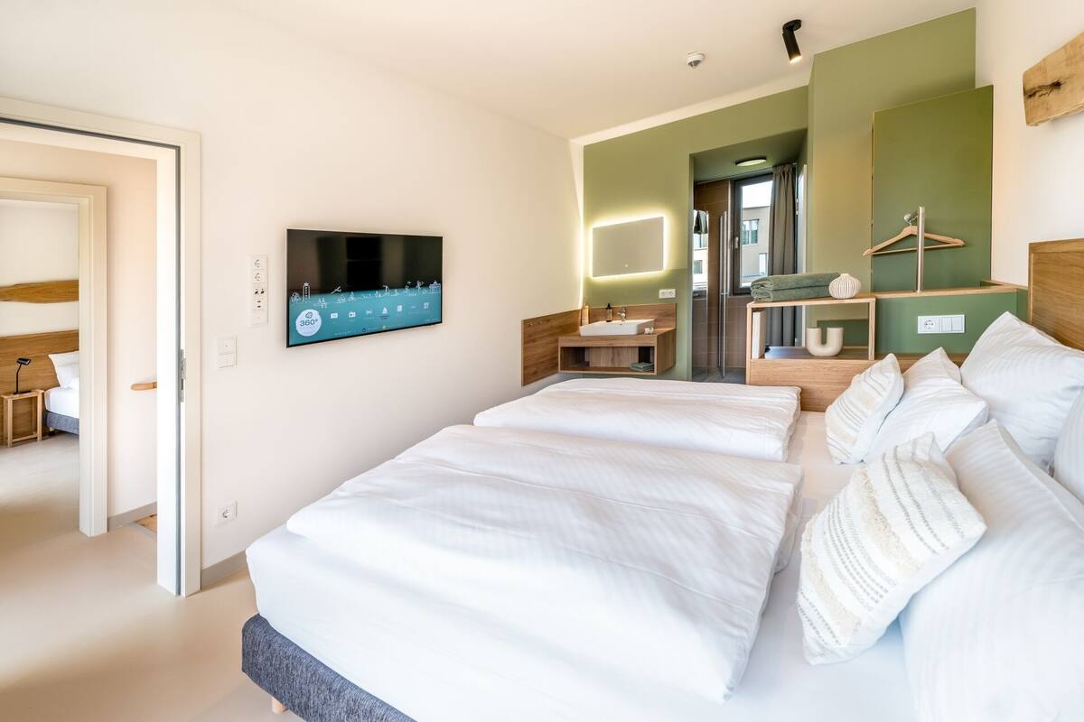 Habitación de hotel moderna con una cama grande, un televisor montado en la pared, un baño con espejo y un cabecero de madera.