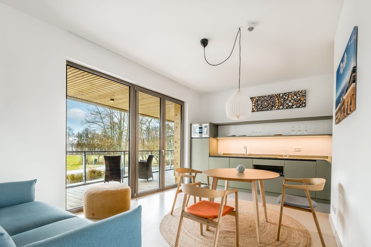 Una sala de estar moderna con un amplio ventanal corredero que da a un balcón, una zona de comedor con una mesa redonda y una cocina equipada con horno y microondas.
