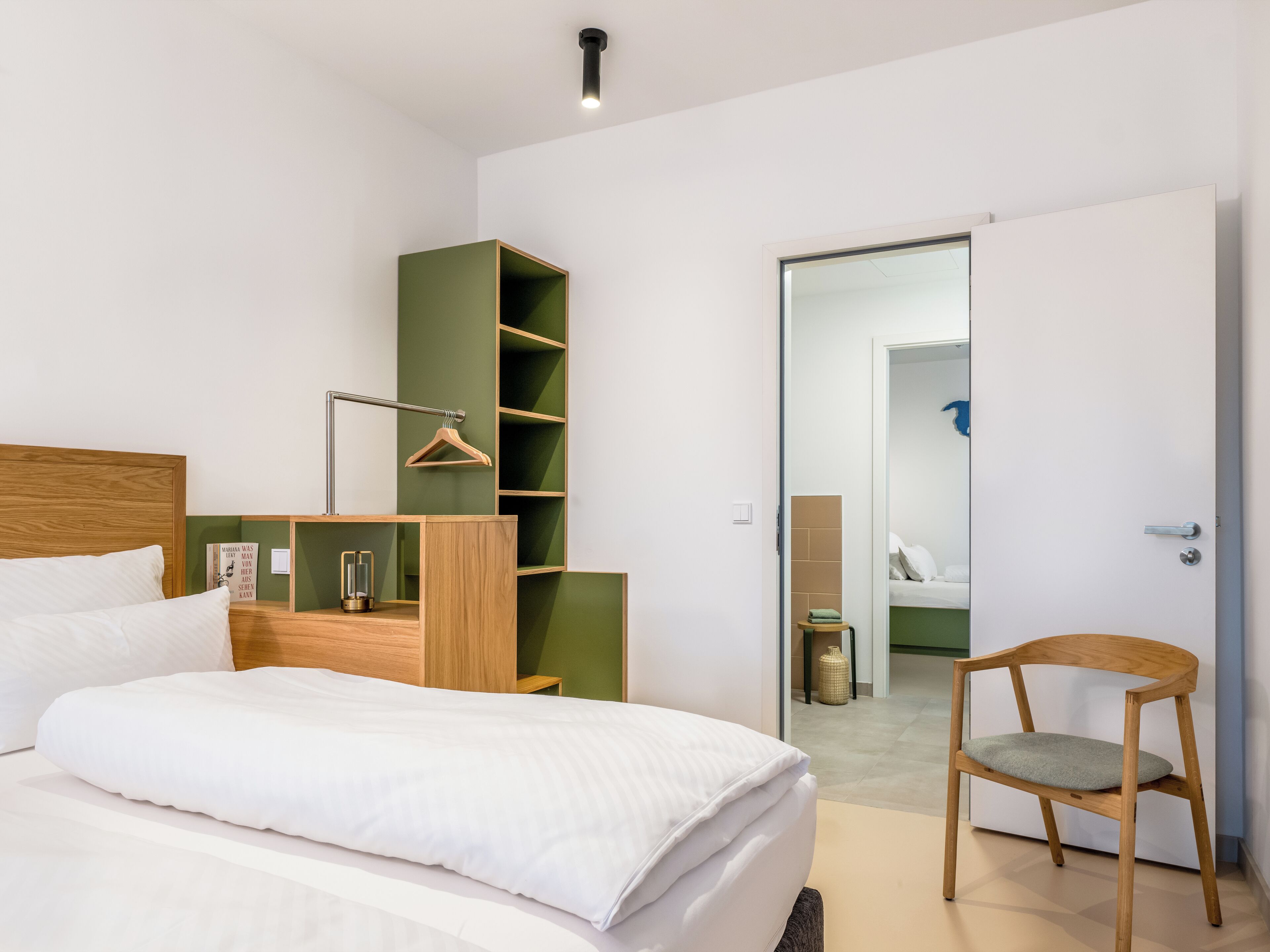 Ein modernes Schlafzimmer mit einem Bett, einem Holzstuhl, einer grünen Regaleinheit und einer Tür zu einem anderen Raum.
