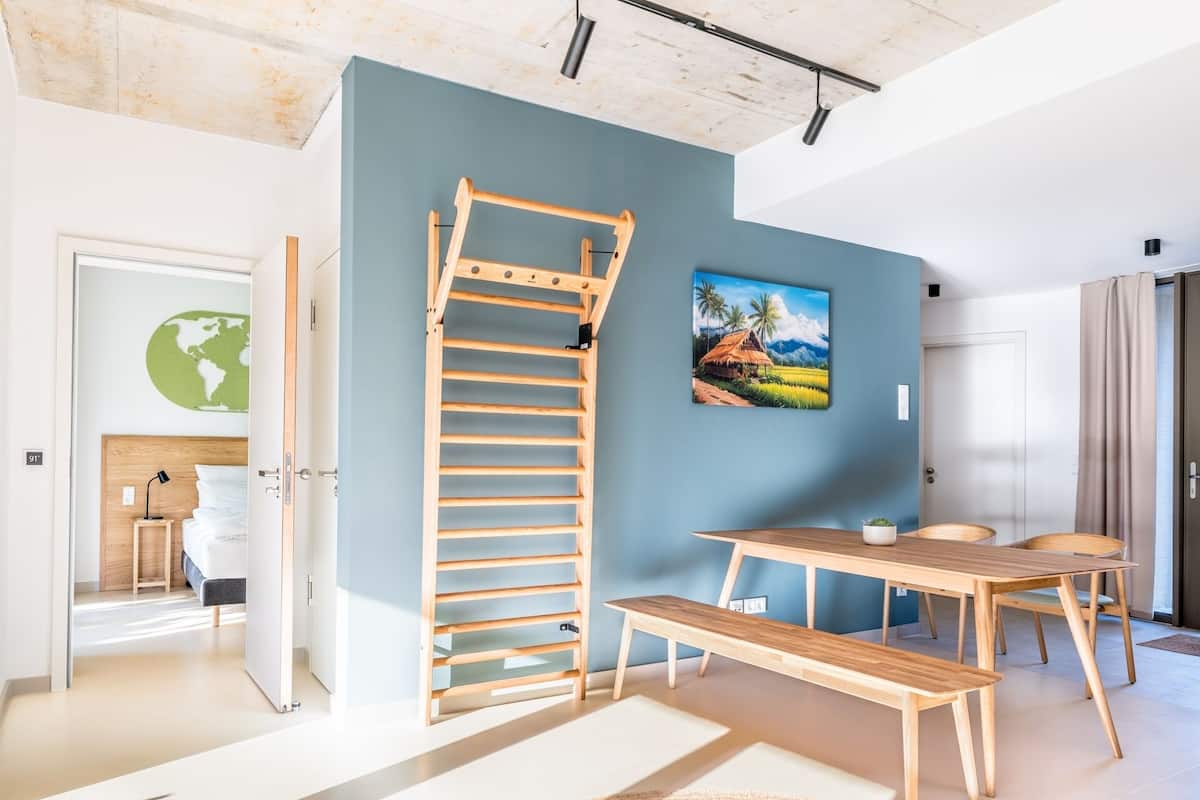 Una habitación con una escalera de madera, una mesa de comedor con sillas, un cuadro de una escena tropical y un globo en la pared.