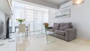 Una sala de estar moderna con una mesa de centro de vidrio, sofá gris, paredes blancas y ventanas grandes.