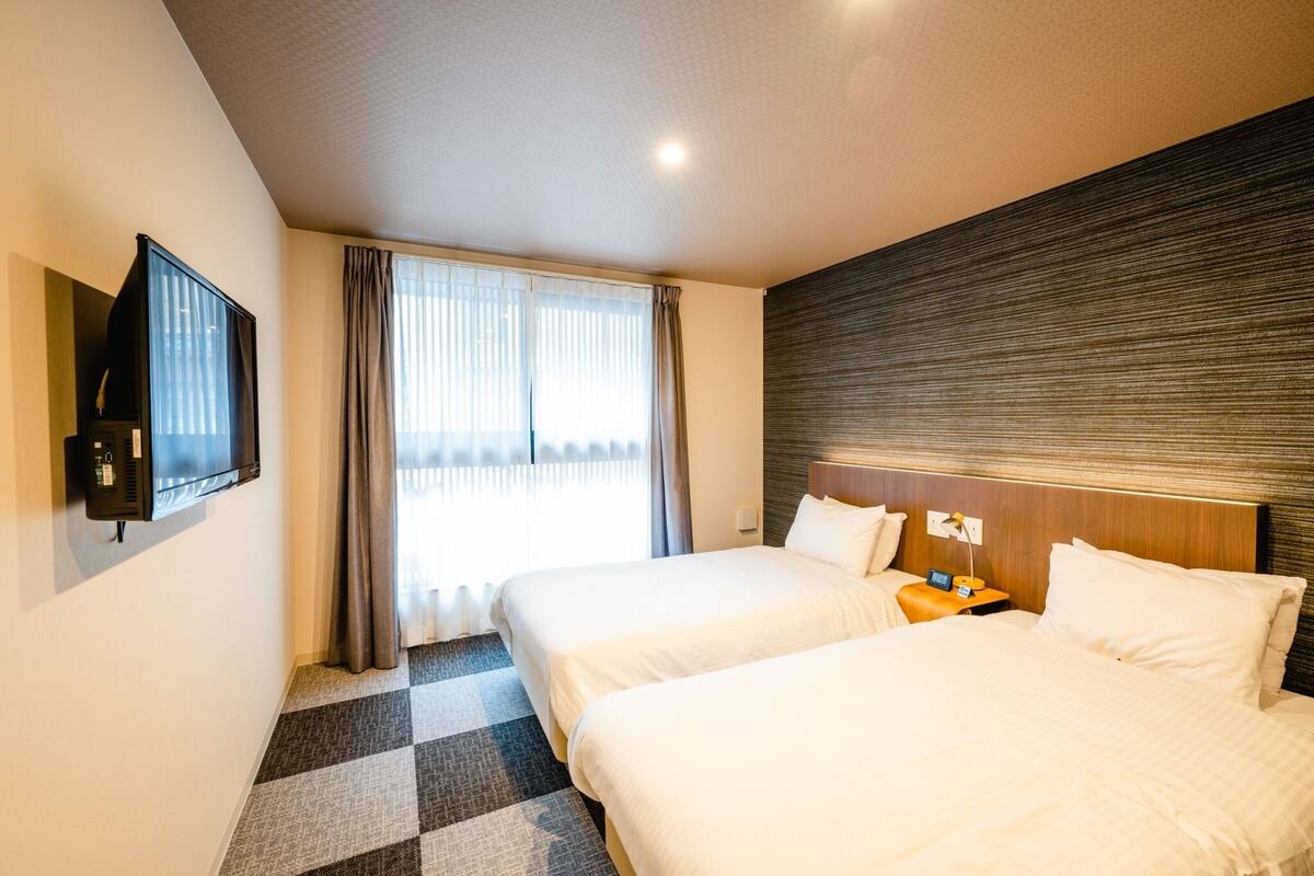 A hotel room with two beds, a flat-screen TV mounted on the wall, a large window with curtains, and a check-in desk.