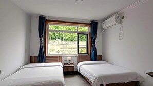 Une chambre d’hôtel avec deux lits simples, une fenêtre avec des rideaux, une table de chevet avec une lampe et un appareil de climatisation fixé au mur.