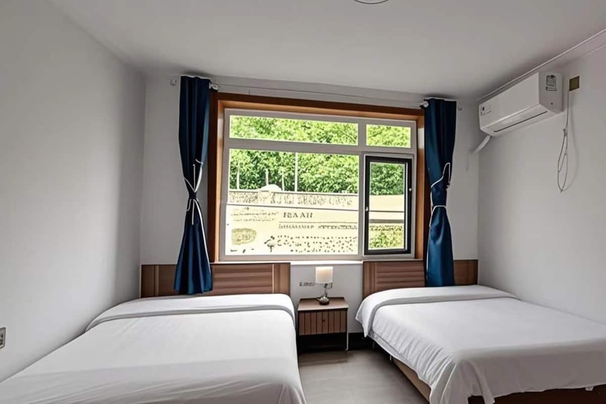 Habitació d'hotel amb dos llits individuals, una finestra amb cortines, una tauleta de nit amb una làmpada i un aparell d'aire condicionat a la paret.