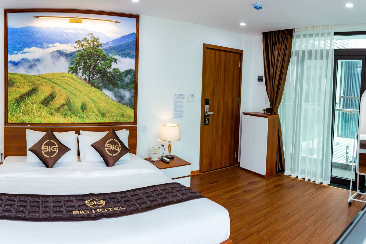 Une chambre d’hôtel avec un grand lit, deux tables de chevet avec des lampes, une tête de lit en bois, un tableau représentant un paysage, une armoire, une fenêtre avec des rideaux et un lit superposé avec une couverture.