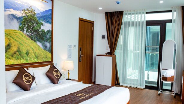Hotelkamer met een groot bed, nachtkastje, bureau, stoel, raam met gordijnen en uitzicht op een bergachtig landschap.