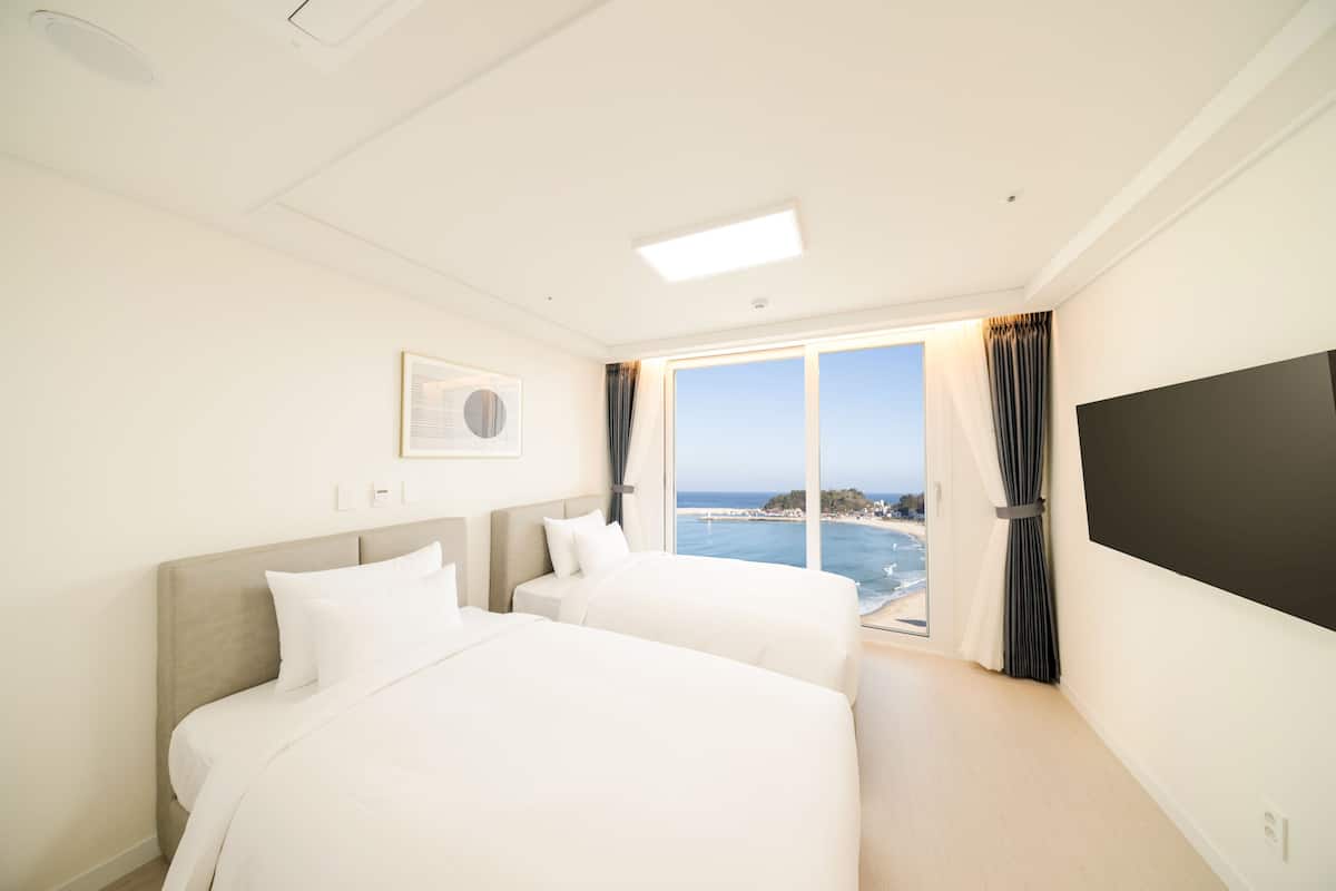 A hotel room with two beds, a large window with a view of the sea, and a flat-screen TV on the wall.