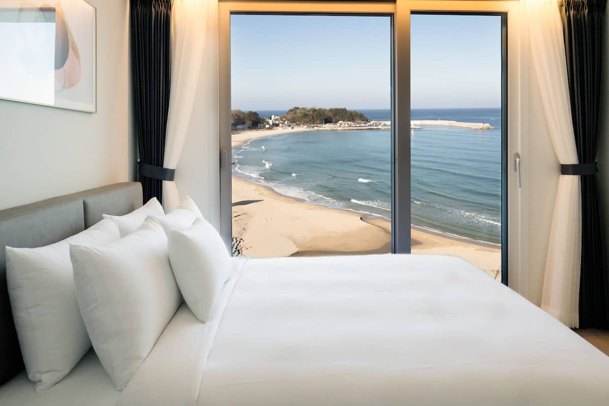 A hotel room with a large bed, white bedding, and a view of the beach and ocean through a glass door.