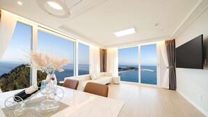A modern hotel room with a large window offering a sea view, a flat-screen TV mounted on the wall, and a dining area with a table and chairs.
