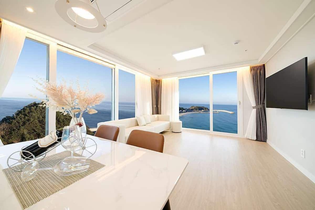 A modern hotel room with a large window offering a sea view, a flat-screen TV mounted on the wall, and a dining area with a table and chairs.