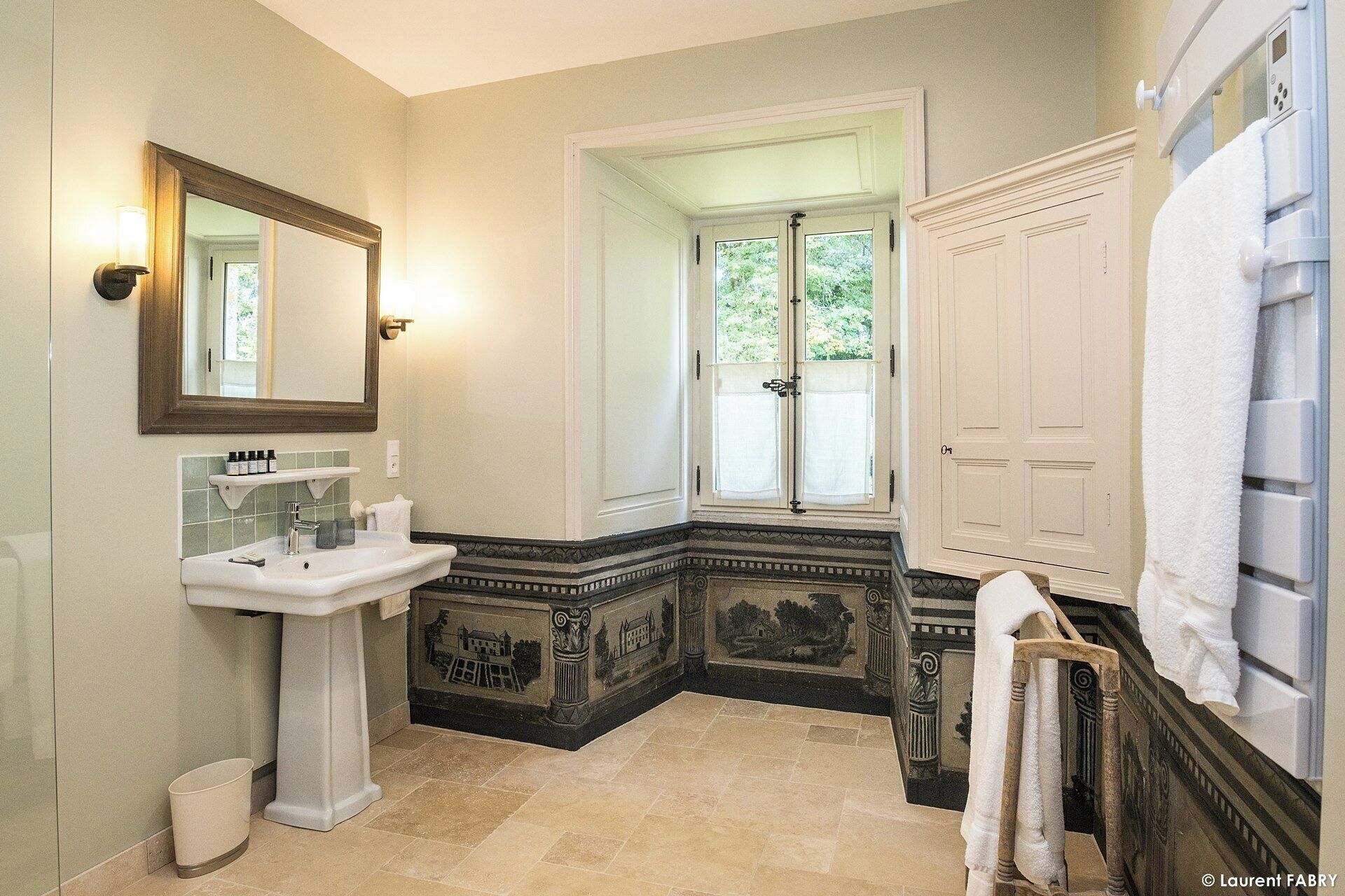 Une salle de bain avec un lavabo sur colonne, un grand miroir, une fenêtre donnant sur un espace verdoyant et une porte blanche.
