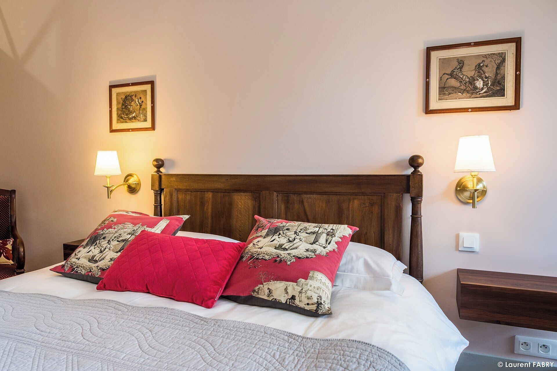 Una cama bien hecha con un edredón acolchado, dos almohadas rojas y un cabecero. Hay dos lámparas fijadas a la pared a cada lado de la cama.
