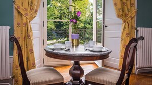 A round dining table with white plates, cups, and a purple vase with flowers, set in a room with yellow curtains and a view of greenery outside. - Chateau de Maillat (Maillat)