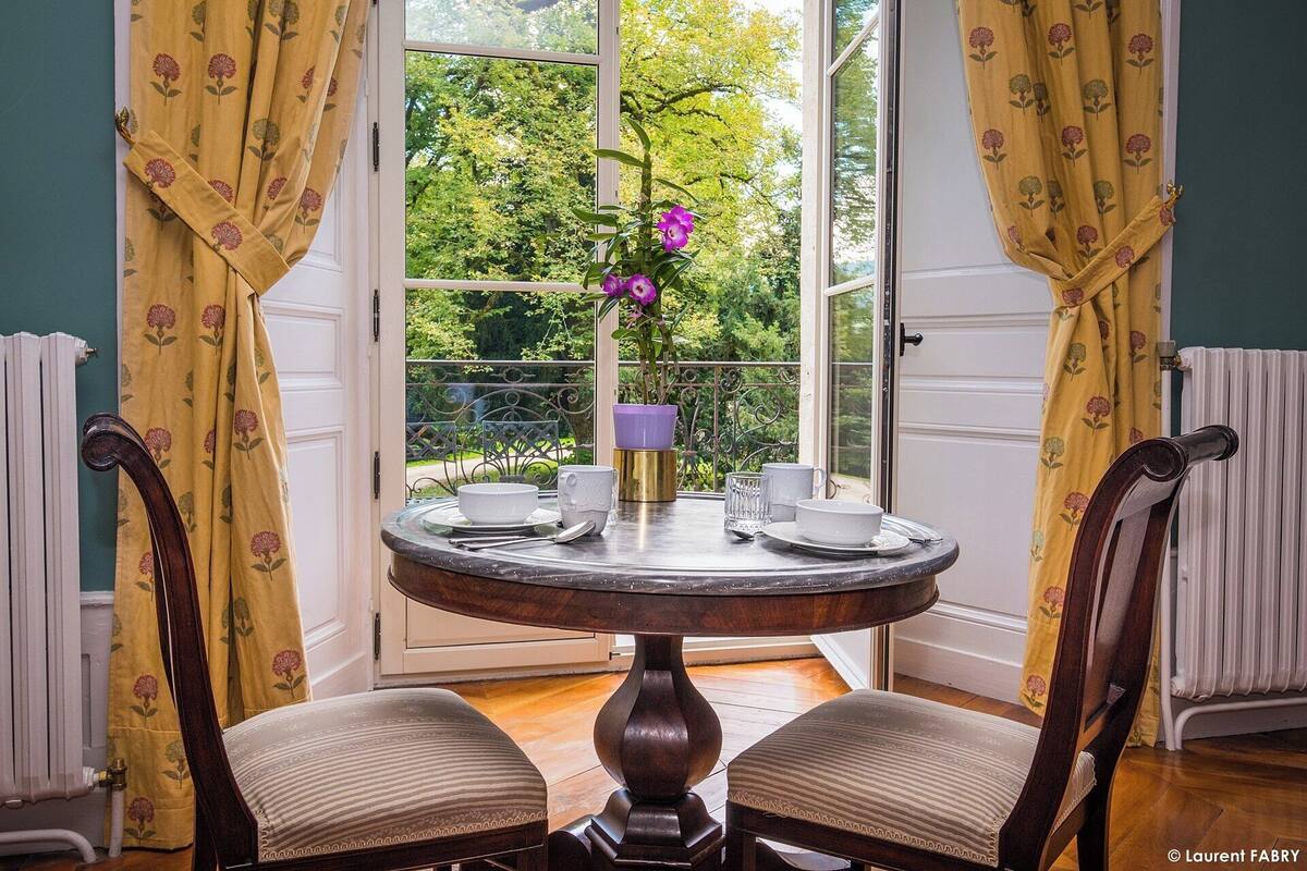 Taula de dinar rodona amb plats blancs, tasses i un vas violeta amb flors, situada en una habitació amb cortines grogues i vistes a la vegetació exterior.
