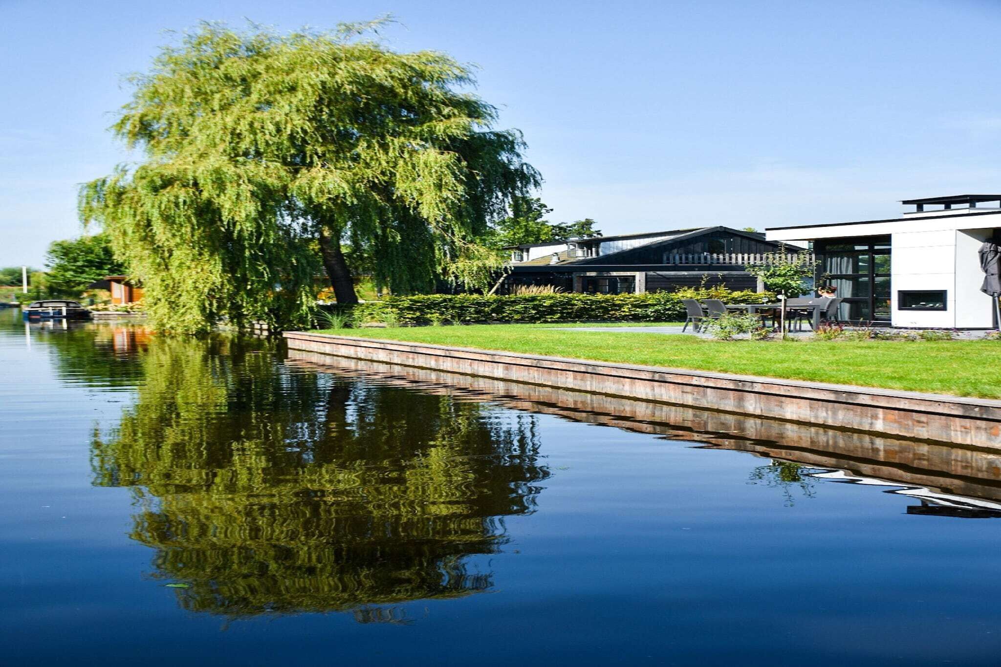 A calm river with a weeping willow tree and a modern building with a glass facade.