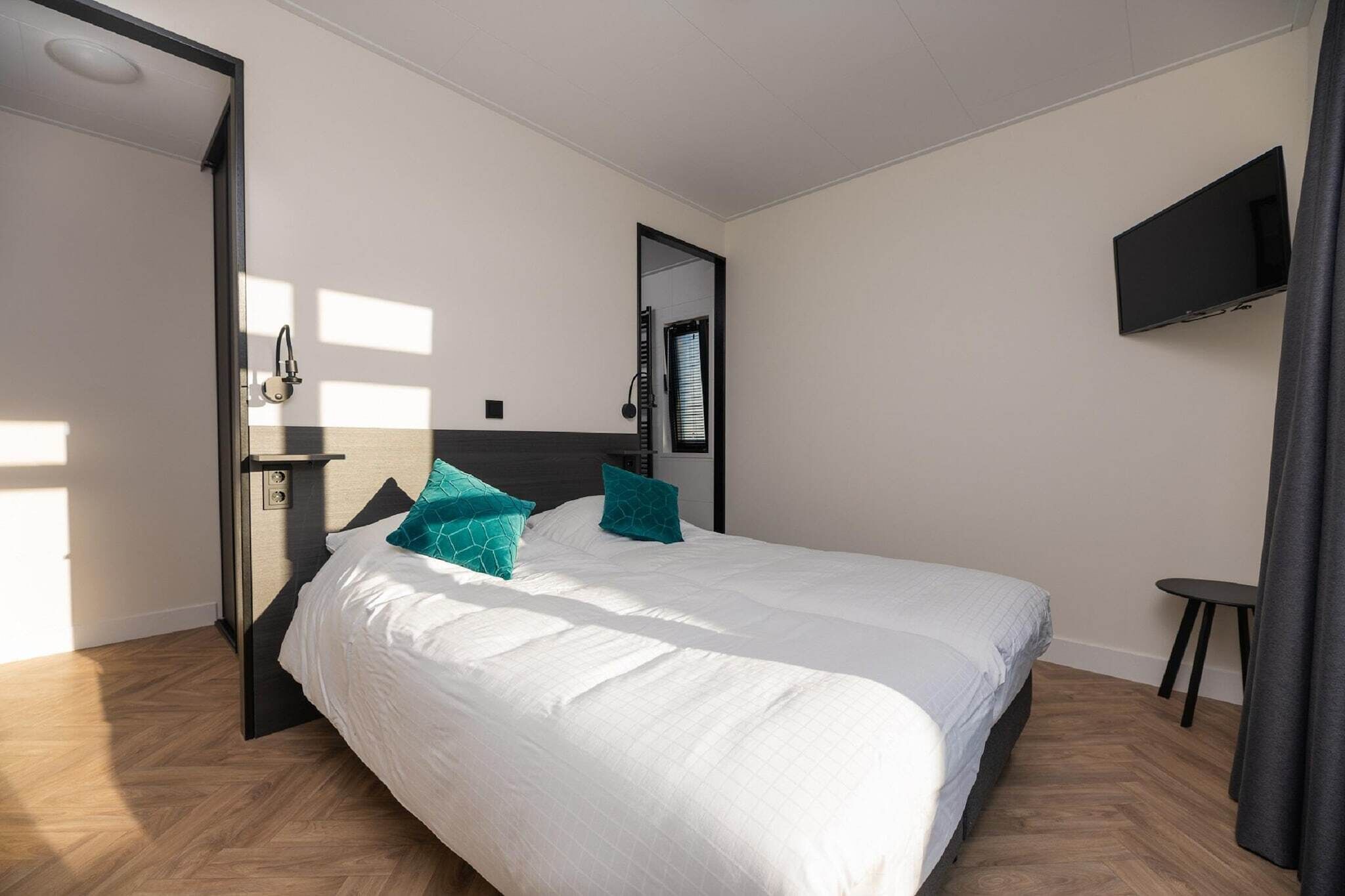Een moderne slaapkamer met een groot bed, een aan de muur bevestigde televisie en een deur naar een andere kamer.