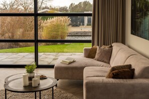 A modern living room with a large window, a beige sofa, a small round table with candles and a book, and a view of a garden and water. - Loosdrecht Water Villa (Nieuw-Loosdrecht)