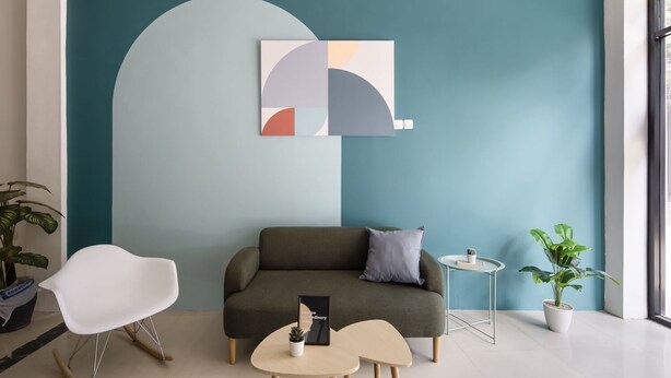 A modern living room with a teal accent wall, a grey sofa, a white chair, and a small round table with a book and a plant. - Cove Veranda (Jakarta)