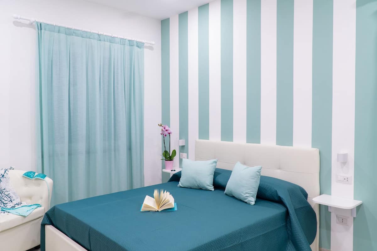 A bedroom with a bed, a nightstand, a potted plant, and a window with curtains.