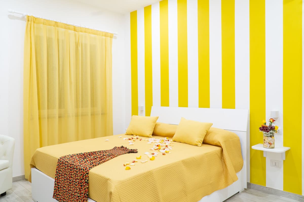 A hotel room with a bed, yellow curtains, a white headboard, and a wall with yellow and white stripes.