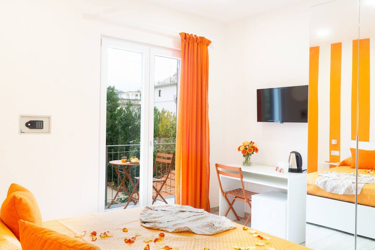 A hotel room with a bed, a desk, a chair, a TV, and a balcony with orange curtains.