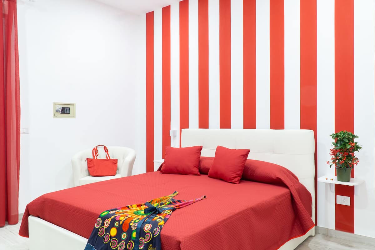 A bedroom with a bed, a chair, a red handbag, a plant, and a wall with vertical stripes.