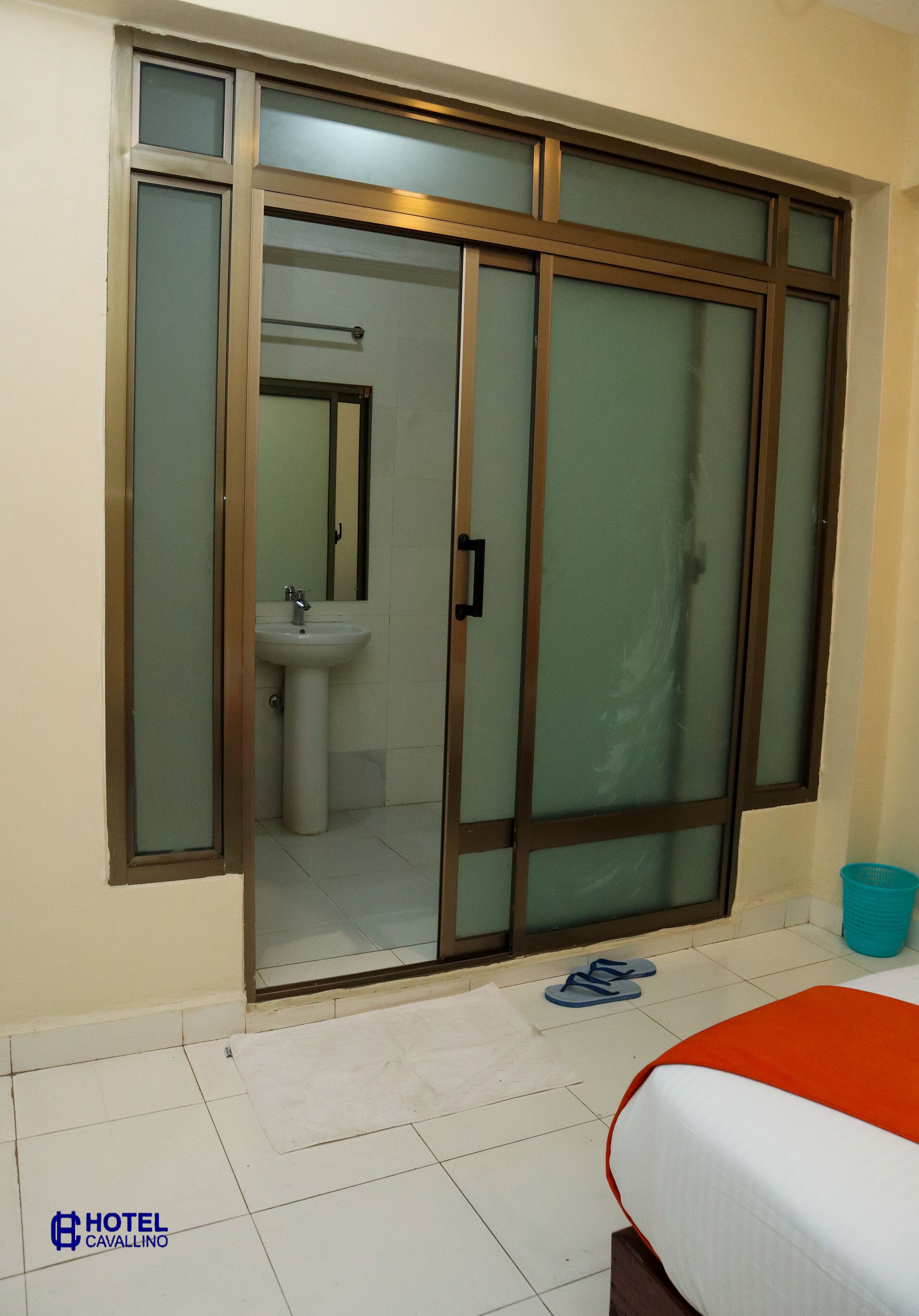 Ein Hotelzimmer mit einer Schiebetür zu einem Badezimmer, einem Bett mit orangefarbener Bettdecke und einer blauen Mülleimer.