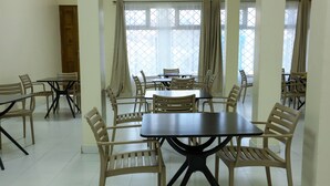 A hotel dining area with multiple tables and chairs, large windows with curtains, and a tiled floor.