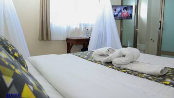 A hotel room with a bed, a TV on the wall, a window with curtains, and a bedside table with a bottle.